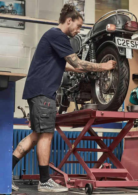 A man in navy blue shorts works on a motorcycle, showcasing rugged cooling stretch cargo shorts with pockets.