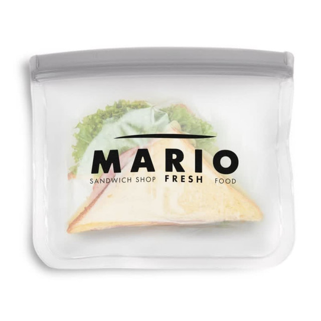 A clear PEVA reusable food storage bag contains a sandwich with lettuce and ham, featuring a logo on the front.