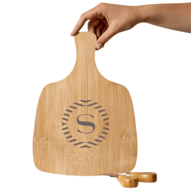 A bamboo cheeseboard with an engraved logo and a hand holding it. Two wooden cheese knives are nearby.
