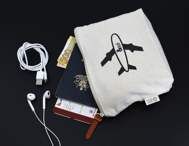 A cream toiletry pouch featuring a logo, containing a passport, currency, earphones, and a charger.