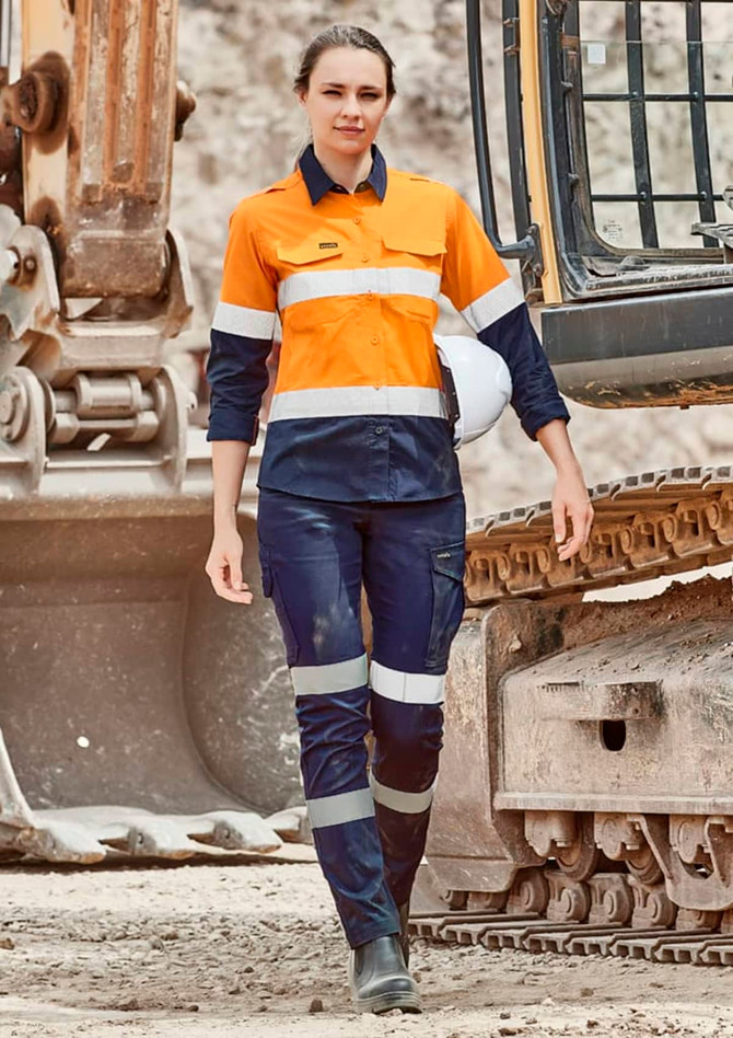 A woman wearing orange and navy cargo pants with reflective stripes, standing near heavy machinery. A hard hat is on her head.