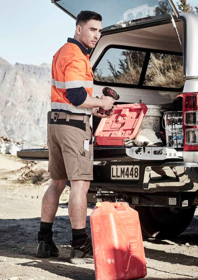 A man in an orange high-visibility shirt and beige shorts stands by an open vehicle boot, holding a tool.