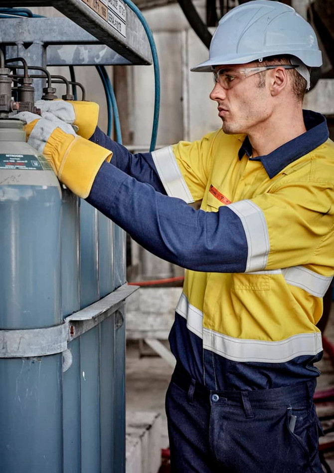 A worker in a yellow and navy spliced shirt adjusts equipment, wearing a hard hat and gloves in an industrial setting.