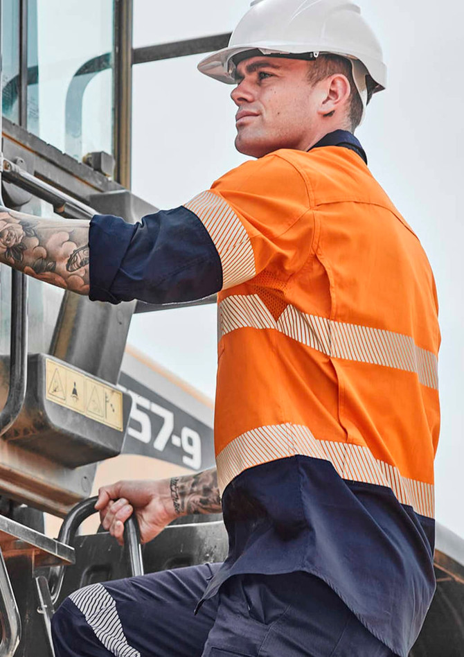 A man in an orange and navy long sleeve hi-vis shirt with segmented tape climbs a machine, wearing a white hard hat.