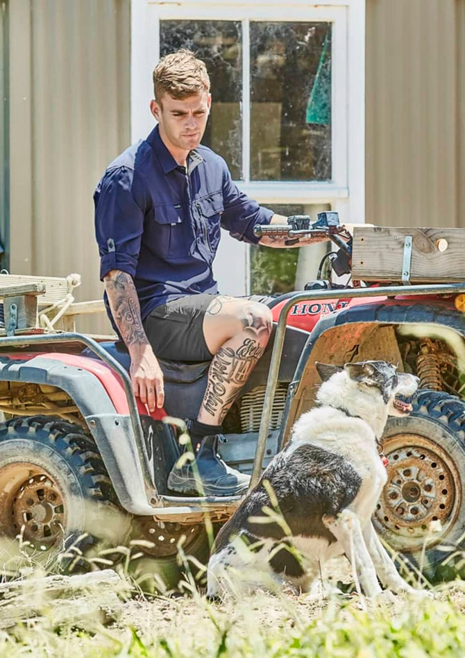 A man in a navy long sleeve button-up shirt sits on an all-terrain vehicle beside a dog. The setting is outdoor with grass.