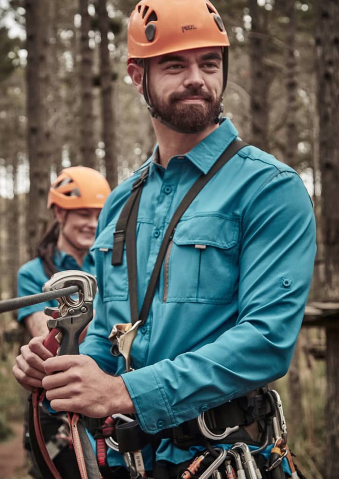 A blue long sleeve button-up shirt worn by a man, in a forest setting, with climbing gear and a helmet.