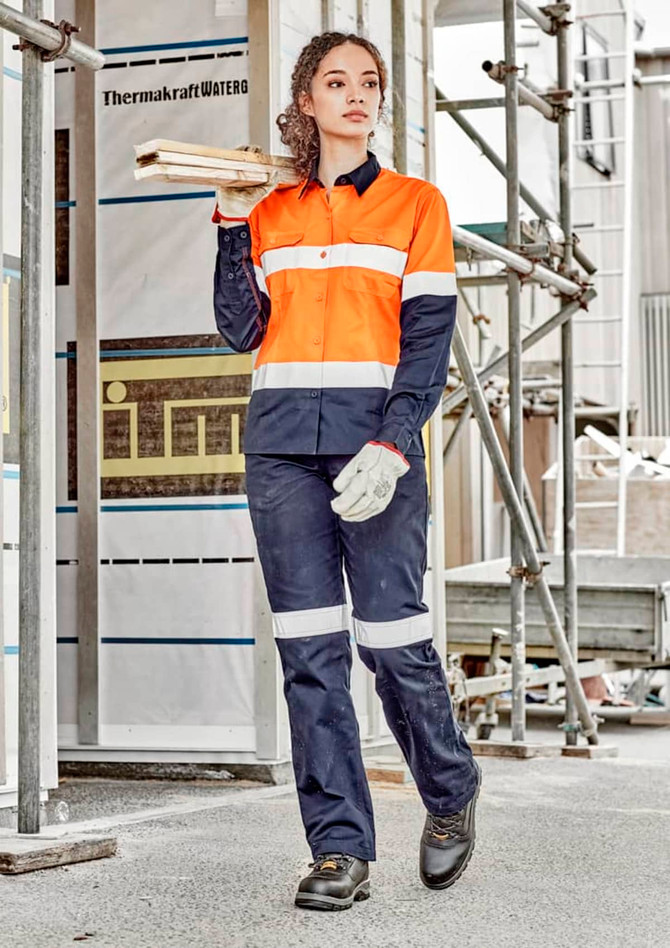 A woman in an orange and navy long sleeve industrial shirt, holding timber, wears work gloves and sturdy boots.