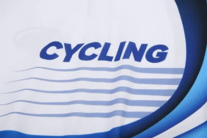 A close-up of a white cycling jersey featuring blue text and wave designs. The jersey includes a logo.