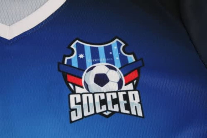 A blue soccer jersey featuring a logo with a shield design and a soccer ball in the centre.