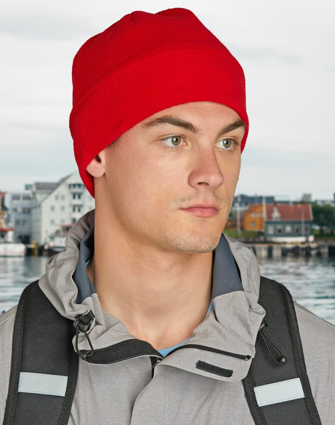 A red beanie worn by a person, paired with a grey jacket, set against a blurred background of a waterfront.