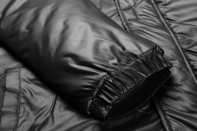 A black Manager Coat Jacket featuring elastic cuffs and a smooth, shiny texture. It has a logo on the fabric.