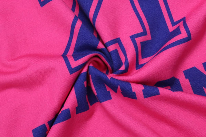 A close-up of a pink cotton hoodie featuring large blue printed letters. The fabric is soft and twisted in the centre.