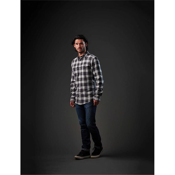 A man stands wearing a black and white checked button-up shirt, paired with jeans and casual shoes.