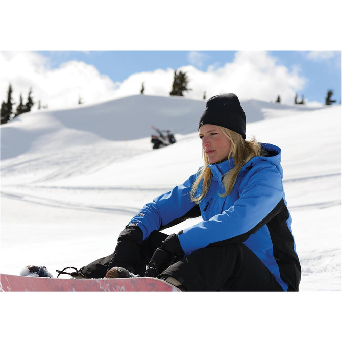 A woman in a blue jacket and black trousers sits on a snowboard in a snowy landscape, wearing a black beanie.