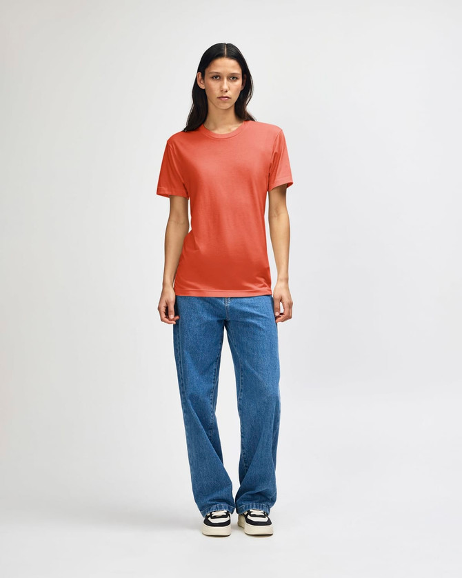 A woman stands wearing an orange American Apparel tri-blend T-shirt and blue denim jeans, featuring a logo.
