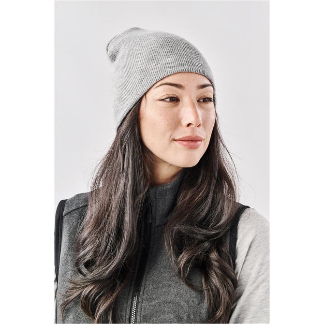 A grey knit beanie worn by a woman with long hair, paired with a grey vest. The beanie has a branding logo.