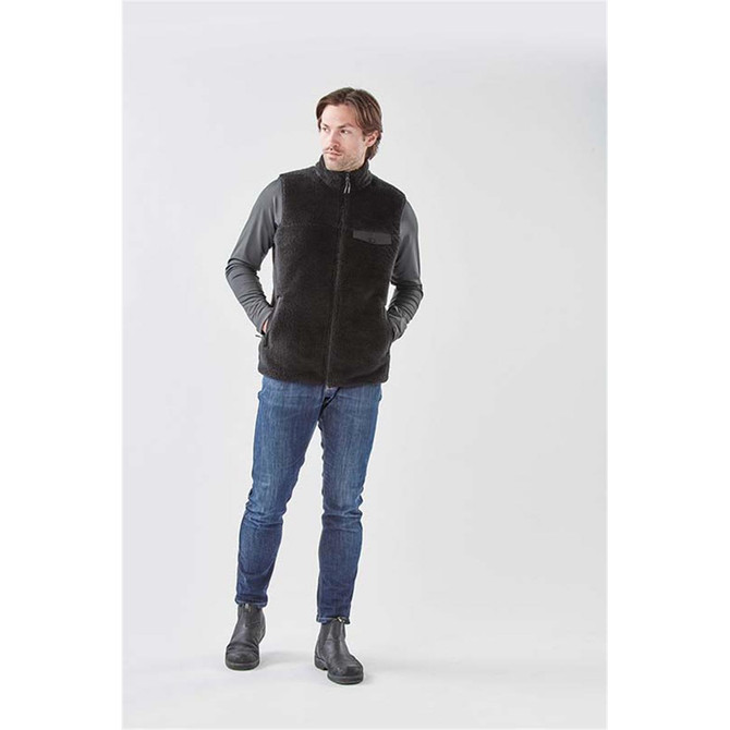 A men's black Sherpa fleece vest, featuring a front zip and pockets, worn over a grey long-sleeve shirt and jeans.