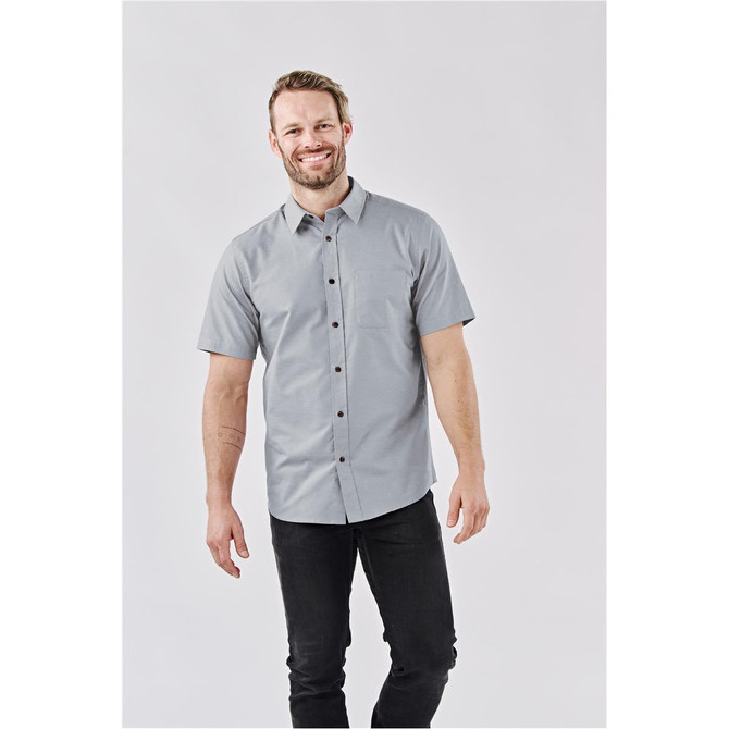 A man wearing a light grey short-sleeve button-up shirt, standing against a neutral background. The shirt features a pocket and a logo.