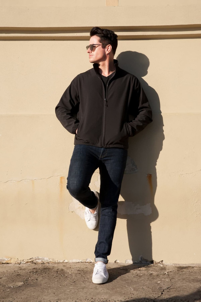 A soft shell jacket in black worn by a man leaning against a wall, paired with dark jeans and white sneakers.
