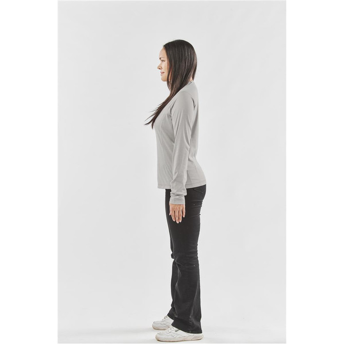 A woman stands in profile wearing a long-sleeve light grey t-shirt and black pants, with a logo visible on the shirt.