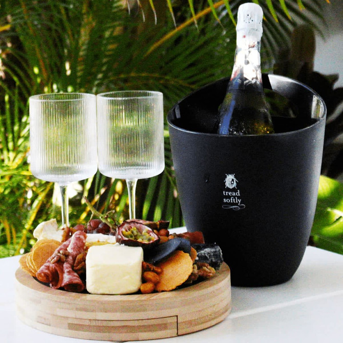 A black ice bucket with a bottle, paired with two clear glasses and a wooden platter of assorted cheeses and charcuterie.