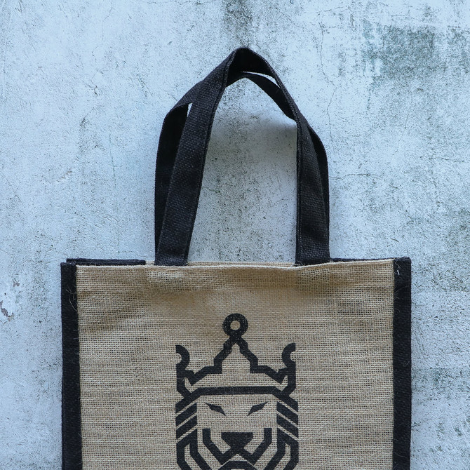 A jute tote bag in natural brown with black handles and a lion design in the centre. It features branding on the side.