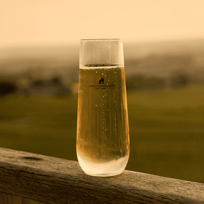 A stemless shatterproof champagne glass filled with golden bubbly liquid, placed on a wooden surface. The glass has a logo.