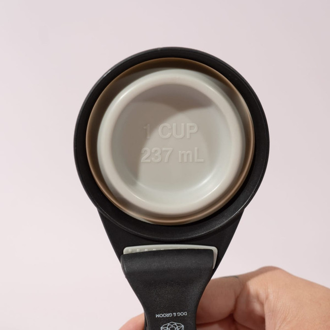 A collapsible silicone pet food scoop in black, featuring a measuring cup with a 1-cup capacity and a bag clip.
