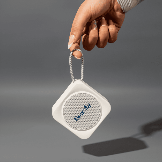 A hand holds a compact white portable Bluetooth speaker with a logo, featuring a loop for easy carrying.