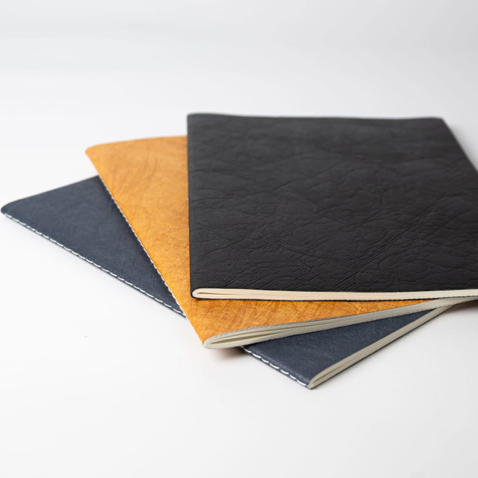 A set of three notebooks in tan, navy blue, and black with textured covers and contrasting stitching.
