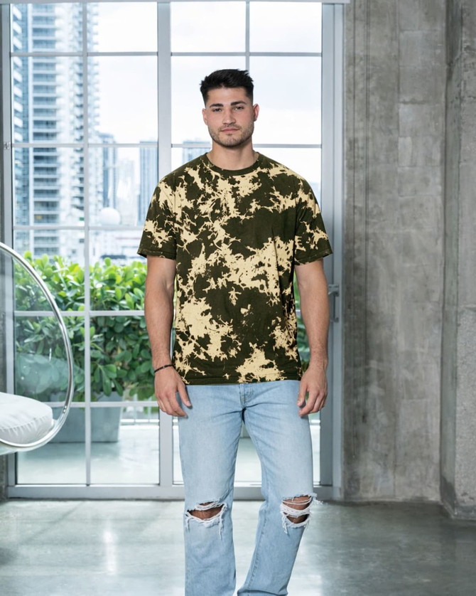 A man stands in a modern interior wearing a bleached tie-dye t-shirt in olive green and cream colours.