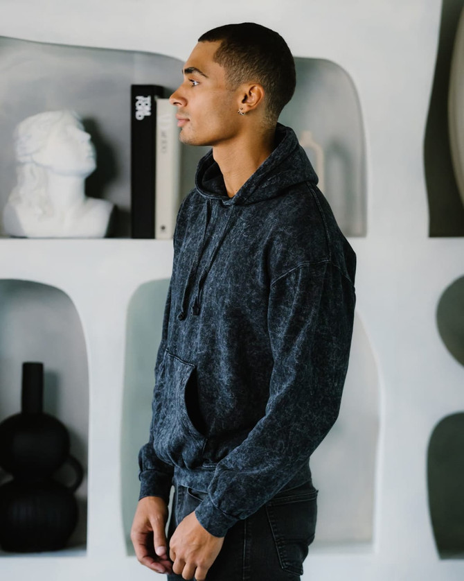 A man wearing a dark grey Colortone® Mineral Wash Pullover Hoodie, standing against a minimalist background.