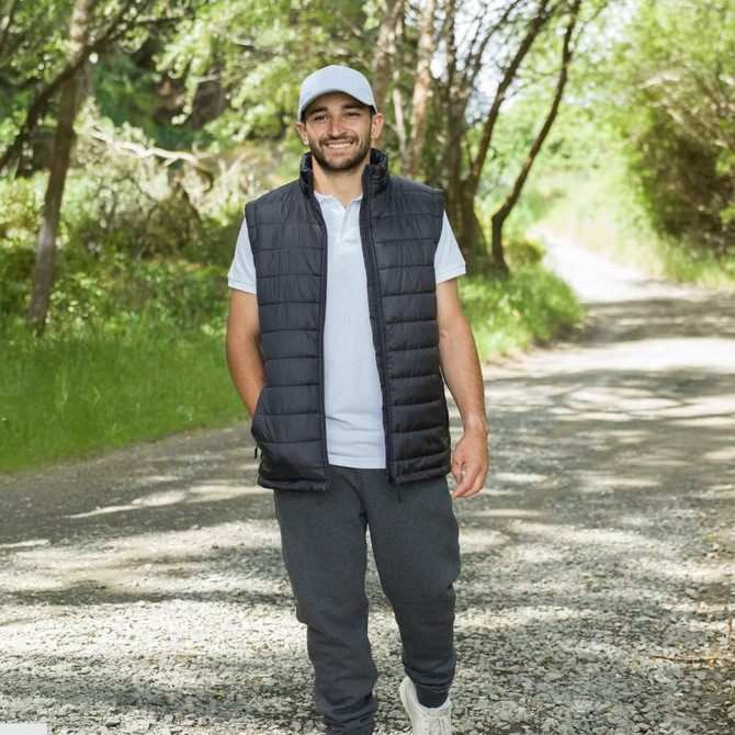 A man wearing a grey polo shirt and a black vest walks along a gravel path surrounded by greenery.