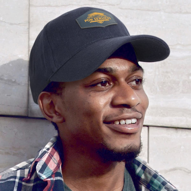 A black cap with a patch, featuring a logo, worn by a smiling man against a textured background.