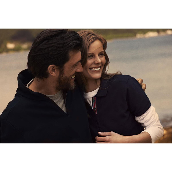 A couple smiles together by the water, wearing dark polo shirts. The woman’s shirt features a subtle striped detail.