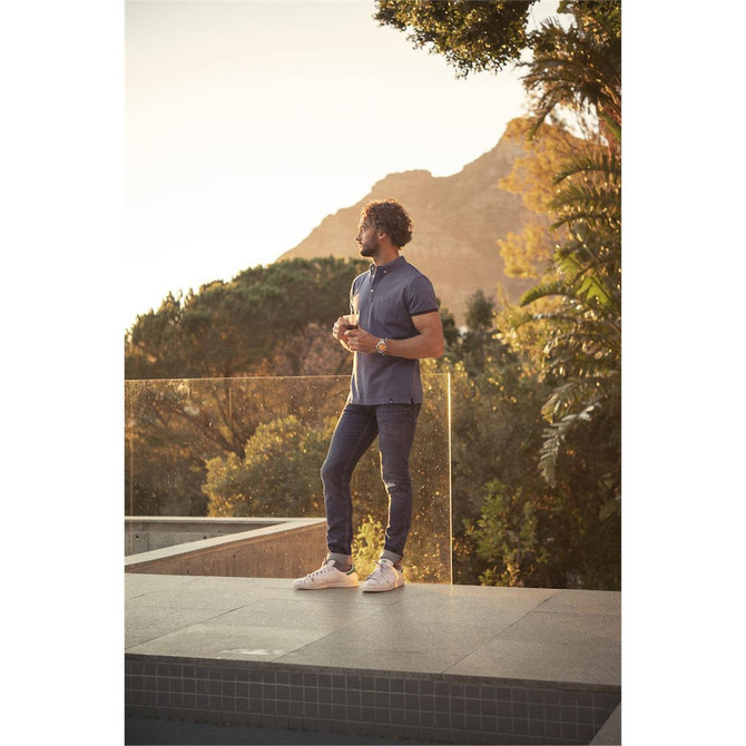 A man stands outdoors in a blue cotton polo shirt and jeans, surrounded by greenery and mountains in the background.