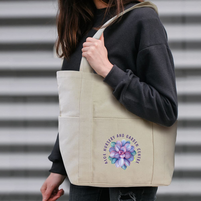 A canvas tote bag in natural colour featuring a floral design and a logo, held by a person wearing a black sweater.