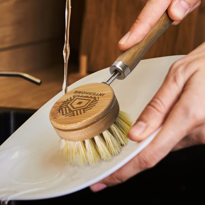 A bamboo dish brush with a wooden handle and natural bristles, being held over a white plate under running water.