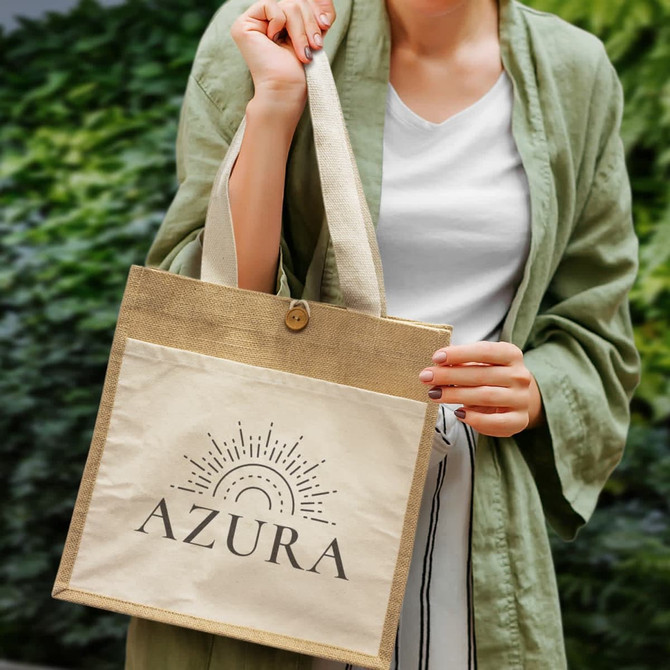 A person holds a beige tote bag with a logo, featuring a jute material base and a cream canvas front.