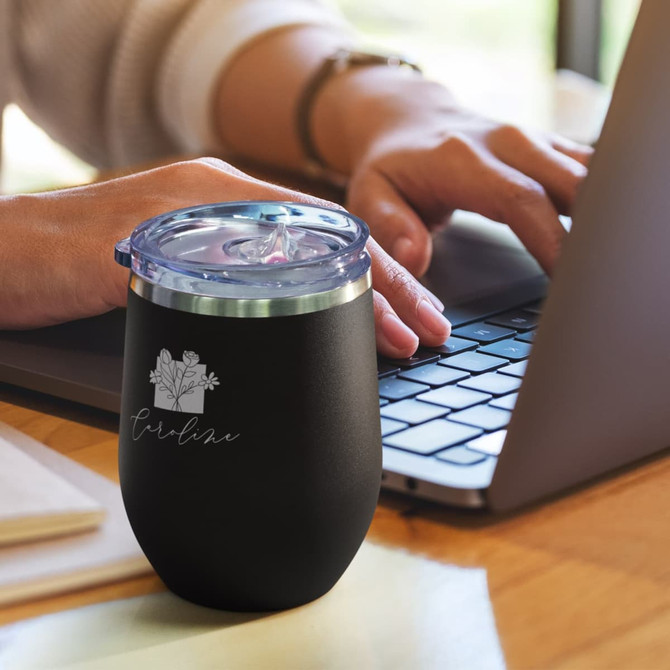 A drink bottle in matte black with a clear lid, placed on a table beside a laptop. It features a logo.