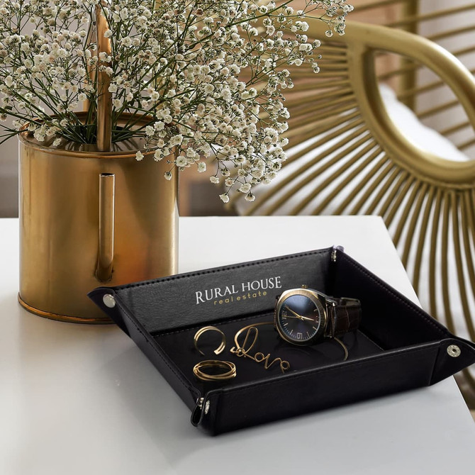 A black valet tray containing a watch and rings, placed on a white table alongside a gold vase with flowers.
