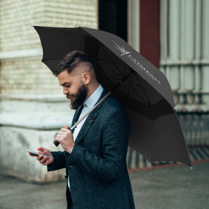 A black urban umbrella held by a man in a suit, standing outdoors while using a smartphone. The umbrella features a logo.