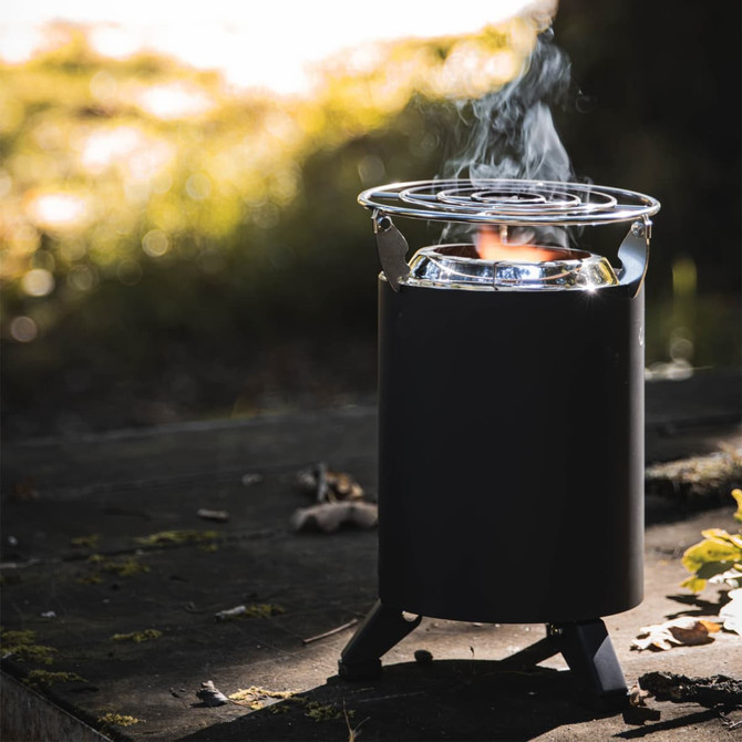 A compact black camp cooker with a grill, producing smoke, set against a natural outdoor backdrop.