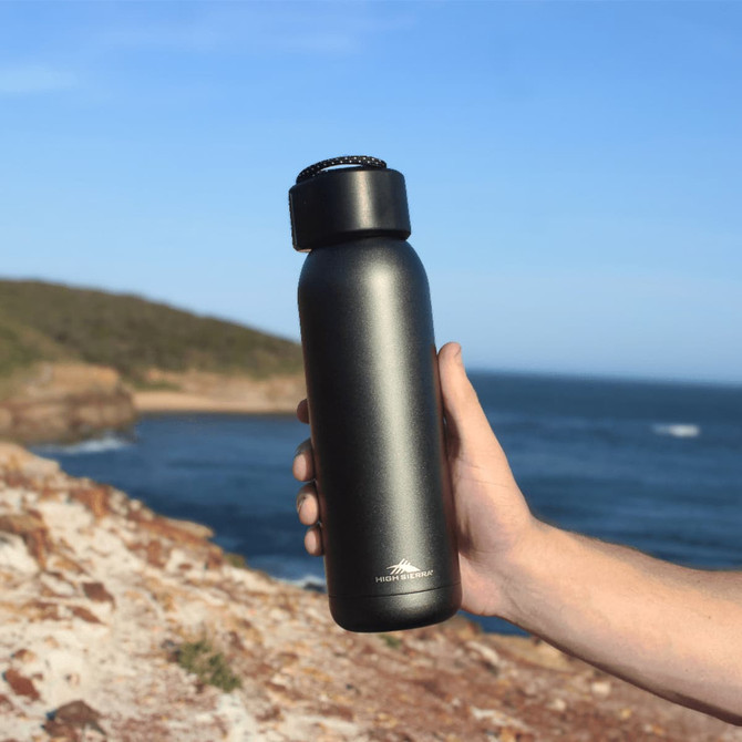 A drink bottle in matte black with a screw top lid, held against a coastal backdrop. Features branding logo.
