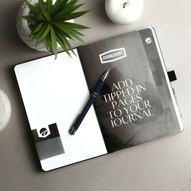 A black and white Nova Bound JournalBook with blank pages, a pen, and a decorative plant on a grey surface.
