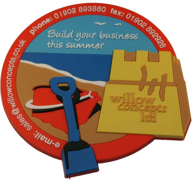 A colourful custom PVC coaster featuring a beach scene, a shovel, and a sandcastle with a logo.