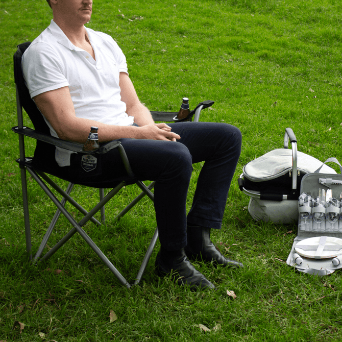 An oversized folding chair in grey, featuring a man seated with a drink. Nearby, a cooler bag and glasses are visible.
