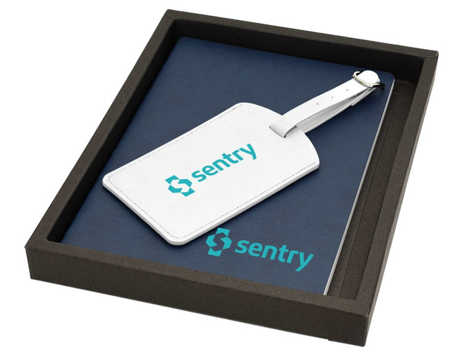 A navy notebook and white luggage tag with a branding logo, presented in a dark box.