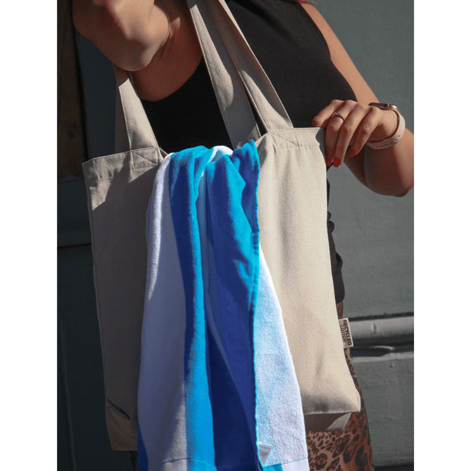 A beige tote bag with a blue and white towel hanging out, held by a person wearing a black top.