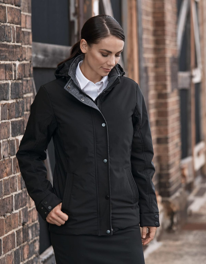 A black jacket with a hood, featuring a front zipper and buttons, worn by a model against a rustic brick backdrop.